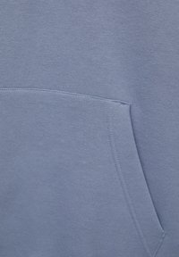 Acercamiento de un bolsillo de tela azul con costuras visibles sobre un material suave y texturizado, probablemente parte de una prenda.