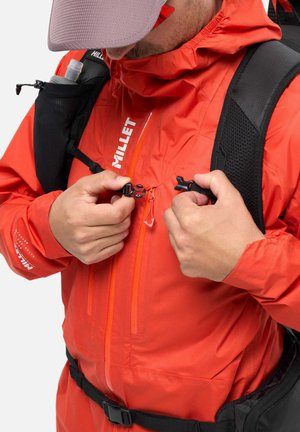 Personne portant une veste Millet orange et une casquette grise en train d'attacher la boucle de poitrine d'un sac à dos noir avec une bouteille d'eau.