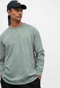Cămașă verde din bumbac cu mânecă lungă, cu decolteu rotund și un mic logo Nike de culoare verde închis pe piept, asortată cu o șapcă neagră.