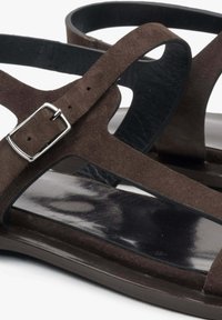 Bruine suède sandalen met een plat ontwerp, een slanke zwarte binnenvoering en een zilveren gespaccent op de verstelbare band.