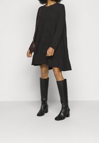 Robe noire à manches longues avec jupons, col rond et texture douce, associée à des bottes noires à talons montant jusqu'au genou et un petit sac bordeaux.