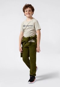 Jauns zēns ar čirkainiem matiem, ģērbies bežā Champion T-kreklā, zaļās sporta biksēs, zaļā džemperī, kas saistīts apvidū, un melnās sporta kurpes, smaida uz priekšu.