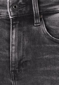 Jeans de mezclilla negra con cierre de botón, costuras distintivas y variaciones sutiles en la textura. Bolsillo visible con un acabado suave.