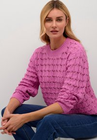 Maglione rosa lavorato a maglia con un motivo aperto testurizzato, scollo e polsini a coste, abbinato a jeans scuri. Il modello ha uno sfondo neutro.