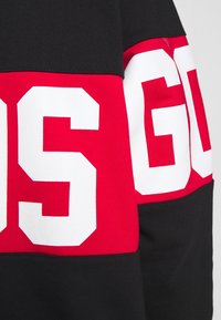 Pullover nero e rosso con lettere bianche audaci. Presenta un design geometrico a blocchi di colore con un tessuto morbido e testurizzato.