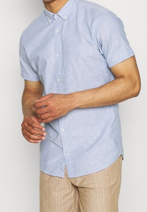 Mann trägt ein hellblaues kurzärmeliges Hemd mit Knöpfen und beige Nadelstreifenhose, richtet seine Hände vor einem weißen Hintergrund.