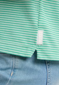 Grün-weiß gestreiftes Hemd aus weichem Stoff, mit einem kleinen, in Weiß gestickten Etikett. Der untere Saum ist an den Seiten etwas länger.