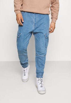 Mand iført lyseblå denim cargobukser med elastiske manchetter og hvide høje sneakers, stående mod en ensfarvet hvid baggrund.