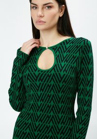 Grüner Strickpullover mit geometrischem schwarzen Muster, langen Ärmeln und einem Schlüsselloch-Ausschnitt mit kleinem silbernen Akzent.