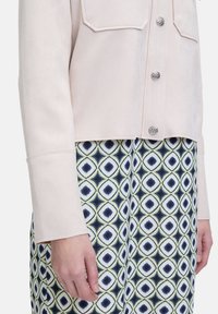 Helles rosa Wildleder-Cropped-Jackett mit sichtbaren Vordertaschen und silbernen Knopfdetails, kombiniert mit einem gemusterten Kleid, das Rautenmuster in Blau und Grün aufweist.