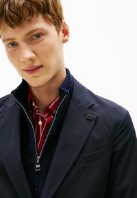 Blazer blu navy in tessuto strutturato, indossato sopra un pile blu navy con zip e una camicia a righe rosse, con un accento a spilla d'argento sul risvolto.