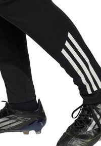 Schwarze Sporthosen mit einem weißen Drei-Streifen-Design an der Seite, kombiniert mit schwarzen Fußballschuhen mit silbernen Akzenten und einer strukturierten Oberseite.