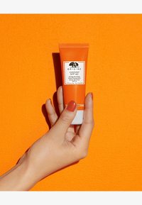 En oransje tube med Origins Ginzing SPF 40 fuktighetskrem holdt i en hånd mot en lys oransje bakgrunn. Etiketten har svart tekst og logo.