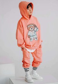 Różowa, oversize'owa bluza z kapturem z grafiką misia, dopasowane spodnie dresowe i białe sneakersy. Posiada zwężane nogawki i skarpetki z logo.
