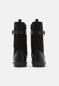 Bottes noires mi-mollet avec bouts de talon en cuir et sangles ajustables, vues de dos sur un fond blanc.