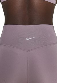 Leggings de sport taille haute en tissu lavande clair, avec une texture lisse, arborant un petit logo blanc de Nike sur la taille.