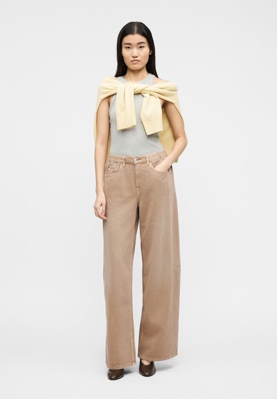 Γυναίκα που στέκεται με το ένα χέρι στην τσέπη, φορώντας μπεζ παντελόνι με φαρδιά πόδια, γκρι αμάνικη μπλούζα, κίτρινο πουλόβερ περασμένο στους ώμους και καφέ παπούτσια.