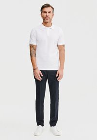 Biała koszulka polo z bawełny, granatowe spodnie i białe sneakersy. Mężczyzna stoi prosto, pokazując tatuaże na ramionach. Prosty, nowoczesny design. Neutralne tło.