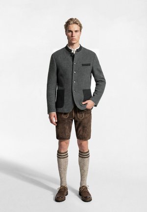 Junger Mann, der eine traditionelle graue Jacke, braune Lederhosen, knielange Wollsocken und braune Lederschuhe vor weißem Hintergrund trägt.