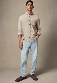 Beige skjorte med oprevne ærmer, parret med lyseblå denim jeans. Mørkebrune sko fuldender outfittet, sat mod en neutral baggrund.