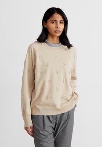Beige pulóver dombornyomott logókkal és kerek nyakkivágással, szürke nadrággal párosítva. Relaxált fazonú, ribbelt mandzsettákkal.