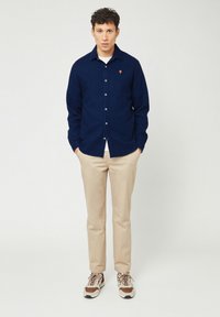 Marineblauwe overhemels met kraag en klein oranje logo, gecombineerd met beige broek en multicolore sneakers. Soepele stoftextuur.