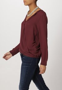 Blusa burdeos de manga larga con cuello en V y diseño drapeado, combinada con jeans oscuros. Un collar de cadena plateada realza el atuendo.
