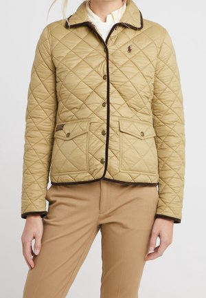 Mujer con chaqueta acolchada beige con ribete oscuro, cierre de botones en la parte delantera, dos bolsillos con solapa al frente y pantalones beige a juego.