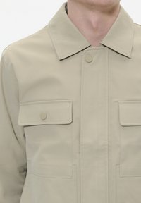 Jachetă bej deschisă dintr-un material texturat. Are un guler ascuțit, două buzunare pe piept și nasturi tip clapetă. Design simplu, utilitar.