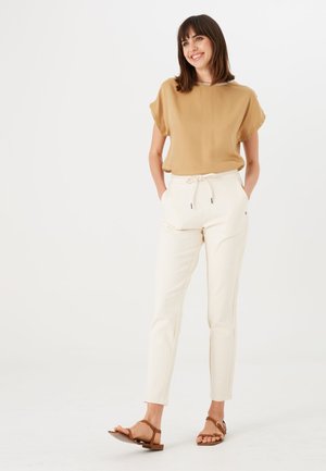 Beige Kurzarmbluse mit rundem Halsausschnitt, kombiniert mit cremefarbenen, taillierten Hosen mit Kordelzug und seitlichen Taschen. Braune Sandalen.