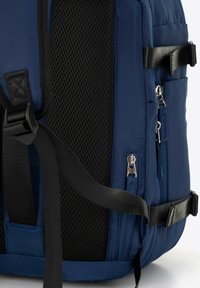 Modrá batoh na boku s černými nastavitelnými popruhy, síťovanou polstrovanou zadní částí a několika stříbrnými zipovými přihrádkami.