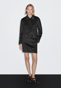 Kvinde iført en mørkegrå dobbeltradet blazer og matchende nederdel, stående foran en ensfarvet hvid væg og sort gulv.