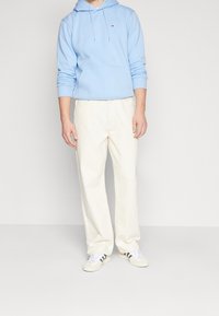Felpa azzurra con cappuccio e tasca frontale, pantaloni color crema e sneakers bianche con strisce nere. Gli abiti hanno una vestibilità comoda e uno stile casual.