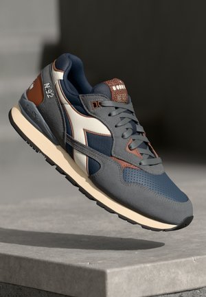 Grey sneaker with navy and white accents, textured upper, cushioned sole, and brown detailing on the heel and tongue; perforated sections.