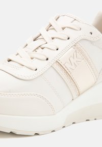 Baskets blanches et beige avec lacets, tissu texturé, surpiqûres lisses et panneau métallique avec le logo "MK" et petits clous sur le côté.