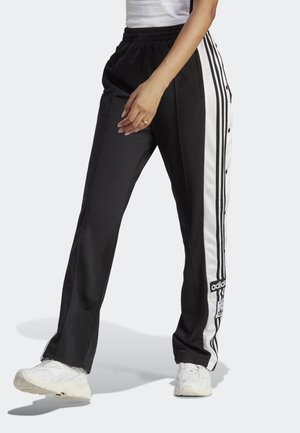 Persona con pantalones de chándal negros de Adidas con paneles laterales y rayas blancas, combinados con zapatillas blancas y una camiseta blanca.