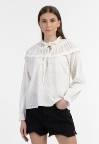 Biała bluzka z falbaniastym kołnierzykiem, związywana na sznurek, z teksturowanym wzorem w kropki. Długie rękawy z elastycznymi mankietami. Połączona z czarnymi szortami.