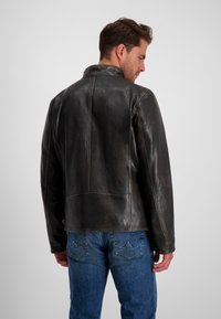 Dunkelbraune Lederjacke mit glatter Oberfläche, ausgestattet mit einem Stehkragen und sichtbaren Nahtdetails. Getragen über blauen Jeans.