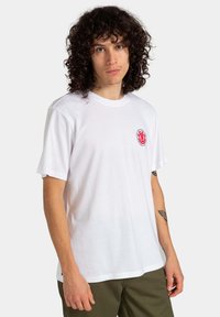 Weißes Baumwoll-T-Shirt mit rundem Ausschnitt und kurzen Ärmeln. Verfügt über ein kreisförmiges rotes und blaues Logo im linken Brustbereich.