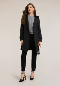 Czarny płaszcz trencz z podwójnym rzędem guzików, dopasowany krój, noszony na teksturowanej szarej bluzce i czarnych spodniach, zestawiony z wysokimi obcasami i kopertówką.