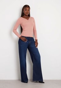 Šviesiai rožinė diržinė ilgų rankovių palaidinė su trimis mygtukais prie kaklo, derinama su tamsiai mėlynomis plačiomis džinsomis ir juodomis smailiomis kulkšnies bateliais.