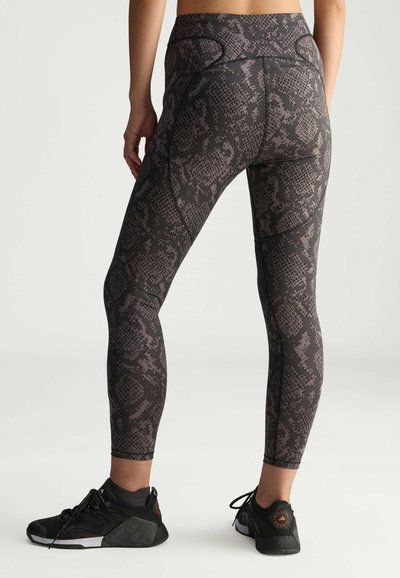 Černé a šedé legíny s hadím vzorem a vysokým pasem, hladkým povrchem a plochými švy. Spárované s černými sportovními botami.