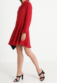 Rotes Kleid mit langen Ärmeln, einem Kragen und schwingendem Rock. Glattes Material, knielang. Schwarze High-Heels-Sandalen mit einem Riemen über dem Fuß.