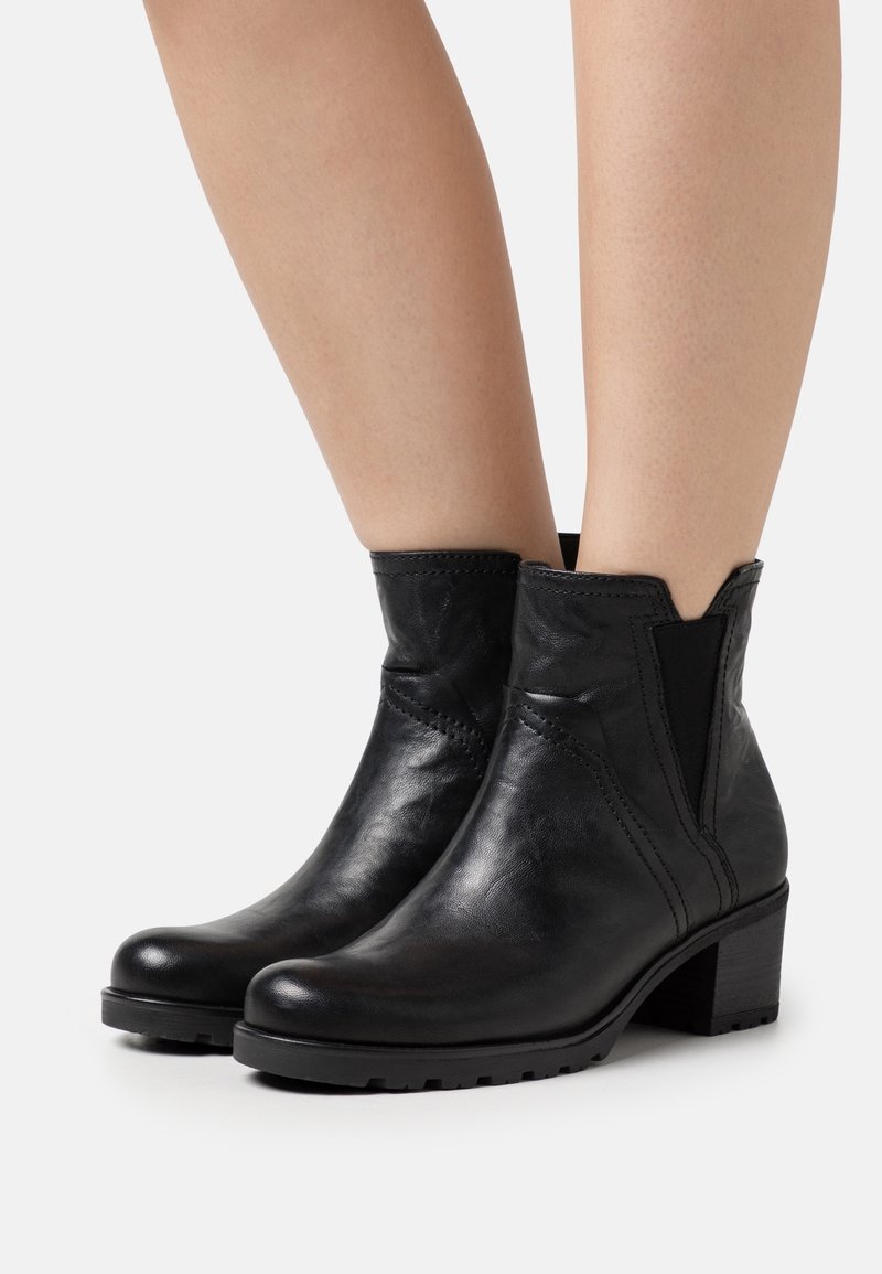 Bottines en cuir noir à talons épais et bas, portées sur jambes nues, avec des panneaux latéraux élastiques et des bouts arrondis.