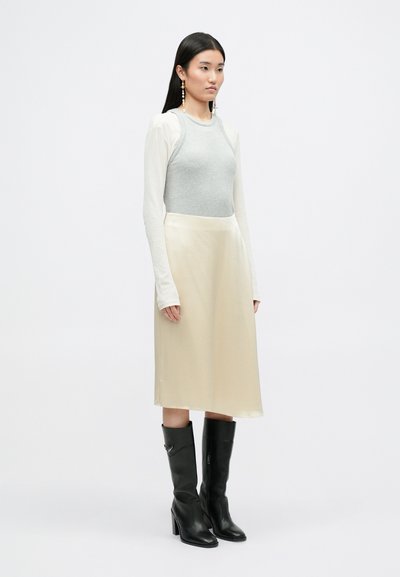 Top gris ajustado de manga larga, falda midi de satén crema y botas de tacón negro hasta la rodilla. Accesorios incluyen pendientes largos. Fondo plano.