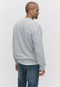 Harmaa collegepaita, jossa on pyöreä pääntie, pitkät hihat, resorit hihansuissa ja helmassa, yhdistettynä sinisiin farkkuihin. Kangas näyttää pehmeältä ja sileältä.