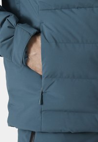 Veste matelassée d'une couleur bleu sarcelle profonde avec une texture lisse ; elle dispose d'une poche latérale et de motifs de couture subtils sur le tissu.