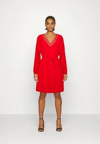 Mulher em pé usando um vestido vermelho até ao joelho com decote em V de renda e cinto na cintura, combinando com saltos altos pretos com tira no tornozelo, sobre fundo simples.