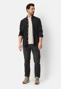Chemise noire boutonnée portée sur un t-shirt crème, associée à un jean foncé et des baskets beiges. Caractéristiques : manches retroussées et coupe décontractée.