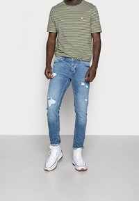 Olivefärgad T-shirt med randmönster, rund hals och en gul logotyp, tillsammans med ljusblå slitna skinny jeans och vita högstövlar.